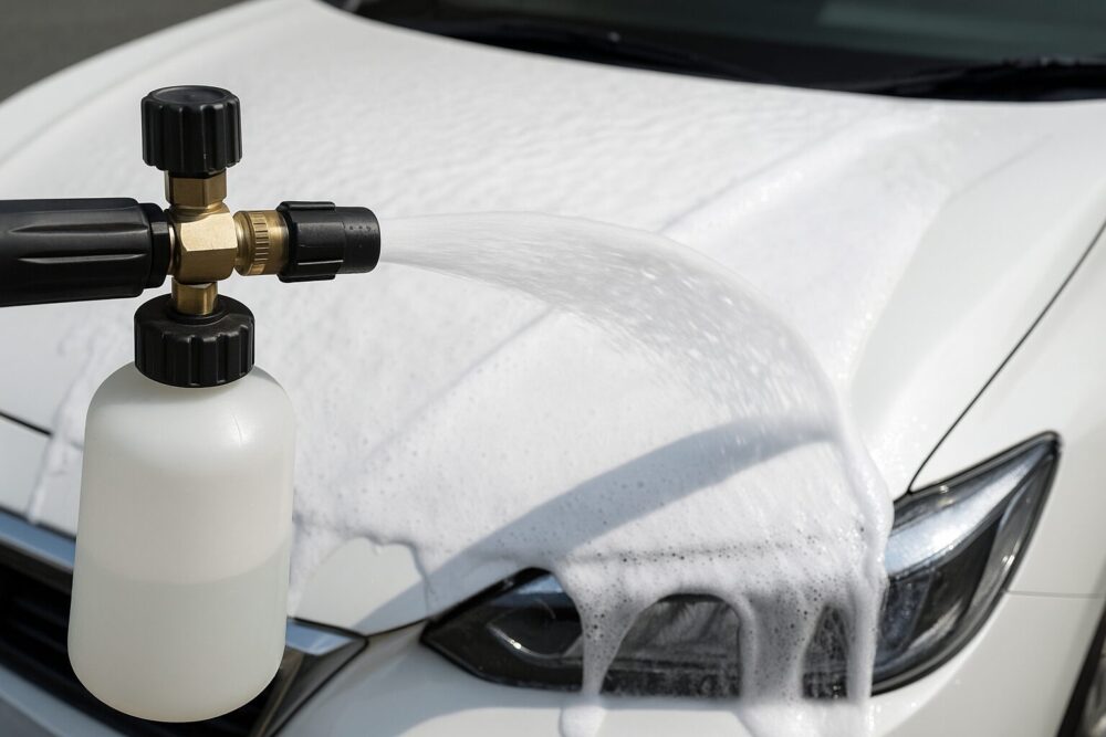 高圧洗浄機ホースに接続したフォームキャノンで、厚い泡の“面”がボンネットを覆う瞬間を接写。クイックカプラと真鍮ノズルが見える角度で、ハイライトは飛ばさず自然な露出。