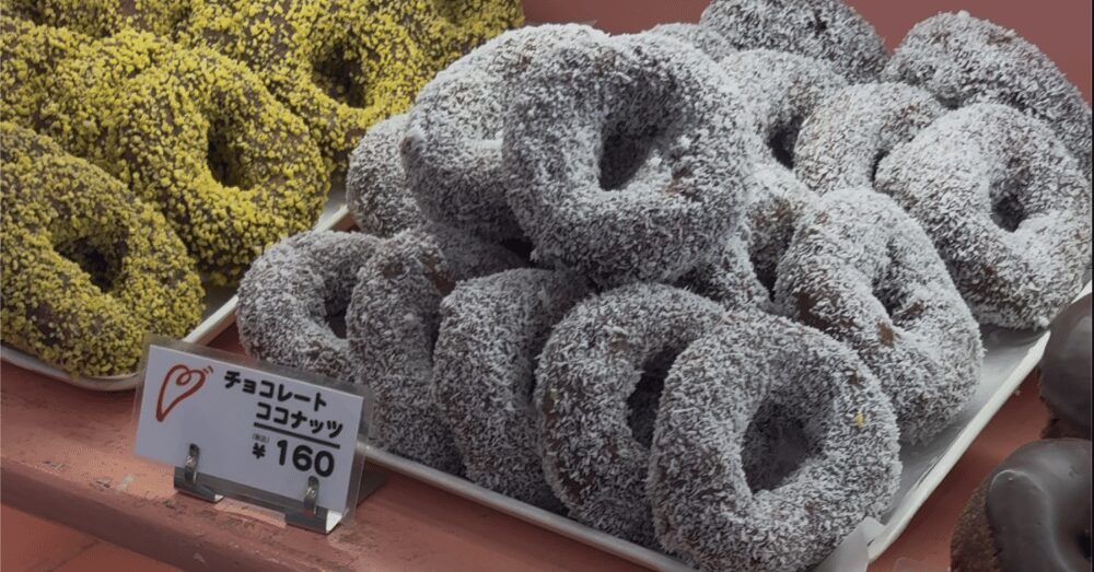 ハッピードーナッツの人気メニュー「チョコレート ココナッツ（税込160円）」。チョコ生地のドーナツに真っ白なココナッツがたっぷりとまぶされ、トレイに山積みになっている様子。