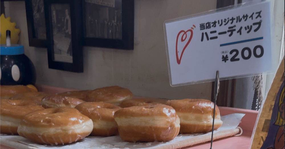 千葉県茂原市のハッピードーナッツ店頭に並ぶ「ハニーディップ」。当店オリジナルサイズ、税込200円と書かれた値札と、ふっくらとした大きなドーナツがトレイに並んでいる様子。背景にはレトロな写真や小物が飾られている。