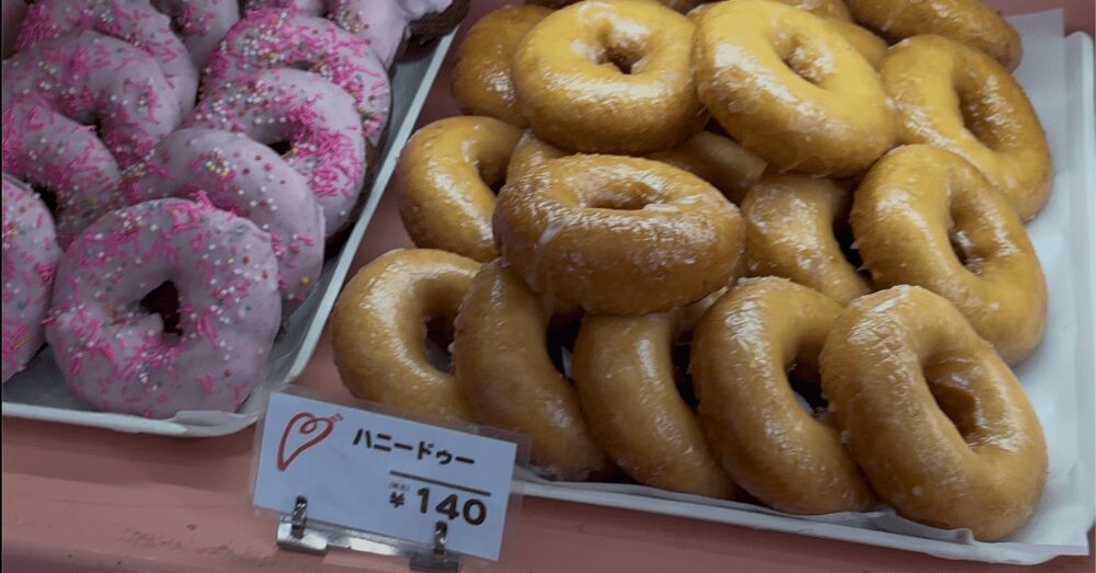 ハッピードーナッツの店頭に並ぶ「ハニードゥー（税込140円）」と、隣に並ぶピンクのスプレーチョコがかかった紫色のドーナツ。山積みになったツヤツヤのハニーグレイスと、ポップなカラーのドーナツが対照的なディスプレイ。