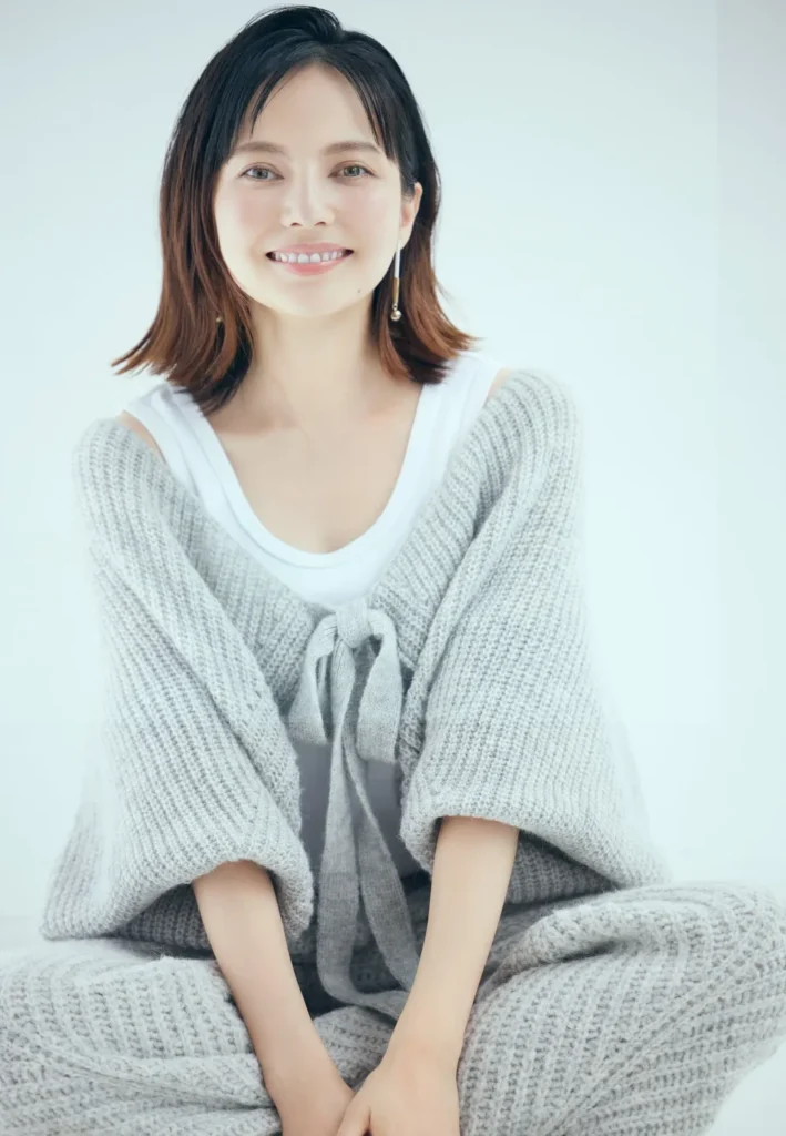 公式プロフィールに掲載されている、グレーのゆったりとしたニットを羽織り、白のインナーを着て笑顔を見せるタレント・ベッキーの顔写真。