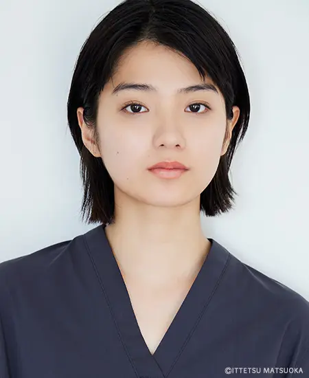 「白バックの前に立つ、黒髪ボブで自然な表情の蒔田彩珠の公式プロフィール写真。ネイビーブルーのVネックを着用。正面からのバストアップショット。」
